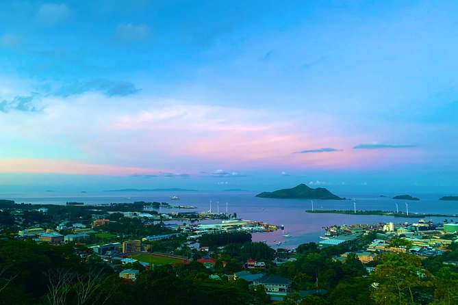 A breathtaking aerial view of a coastal city in Seychelles at sunset, with the sky painted in soft pink, purple, and blue hues. The harbor is dotted with boats, and small lush green islands are visible in the calm waters. The foreground features a mix of colorful buildings and tropical vegetation, while wind turbines stand along the shoreline in the distance.