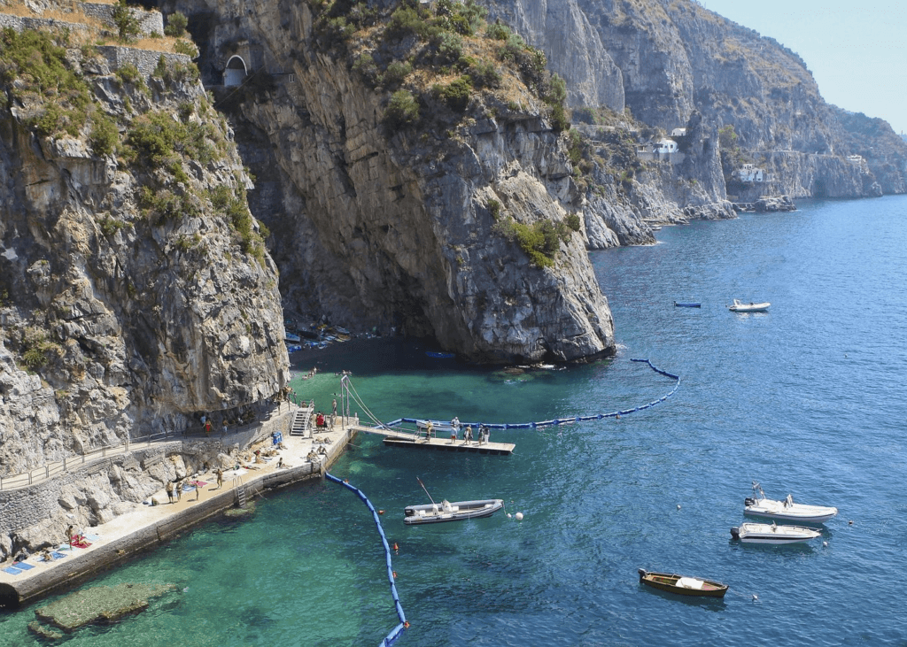 Luxury Amalfi Coast destination featuring a secluded cove with crystal-clear turquoise waters, dramatic limestone cliffs, and elegant boats floating near a sun-soaked beach, offering an exclusive and picturesque Mediterranean escape