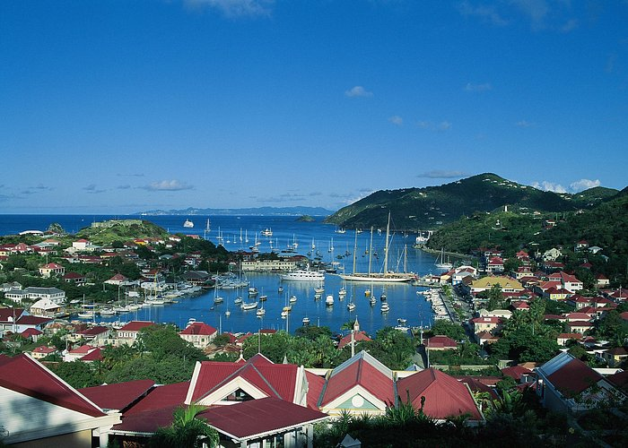 A scenic view of a coastal harbor town with red-roofed buildings nestled among lush green hills. The deep blue waters of the bay are dotted with numerous sailboats and yachts, while a pier extends into the harbor. In the background, small islands and the open sea stretch toward the horizon under a bright blue sky with a few wispy clouds.